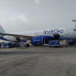A white and blue IndiGo passenger airplane parked on an airport tarmac, viewed from the side under a hazy sky.