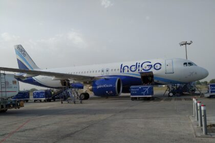 A white and blue IndiGo passenger airplane parked on an airport tarmac, viewed from the side under a hazy sky.