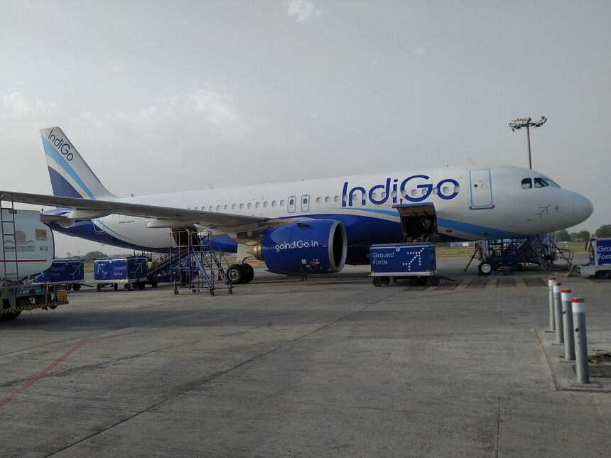 A white and blue IndiGo passenger airplane parked on an airport tarmac, viewed from the side under a hazy sky.
