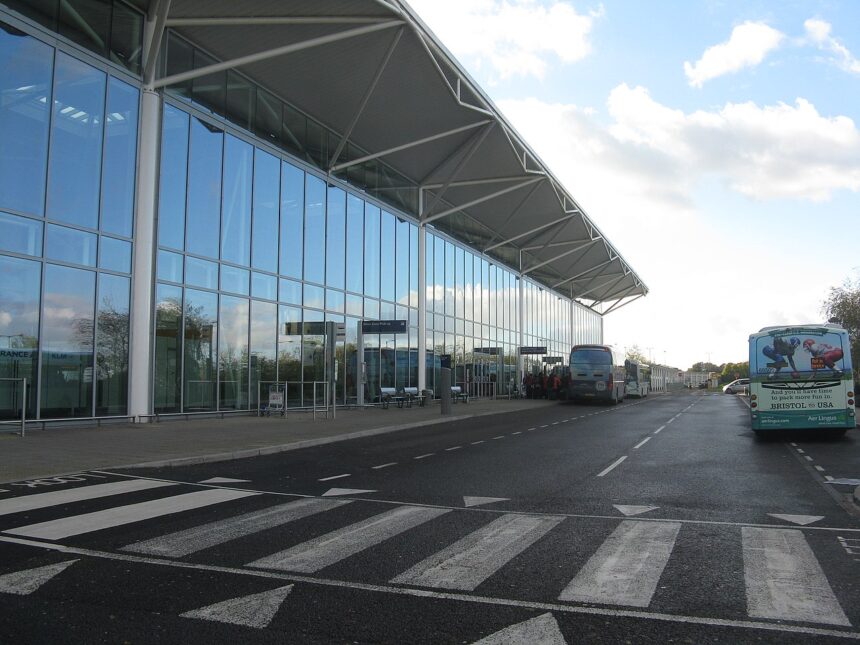 Glass-fronted terminal at Bristol Airport with buses parked outside and a roadway with pedestrian crossing.