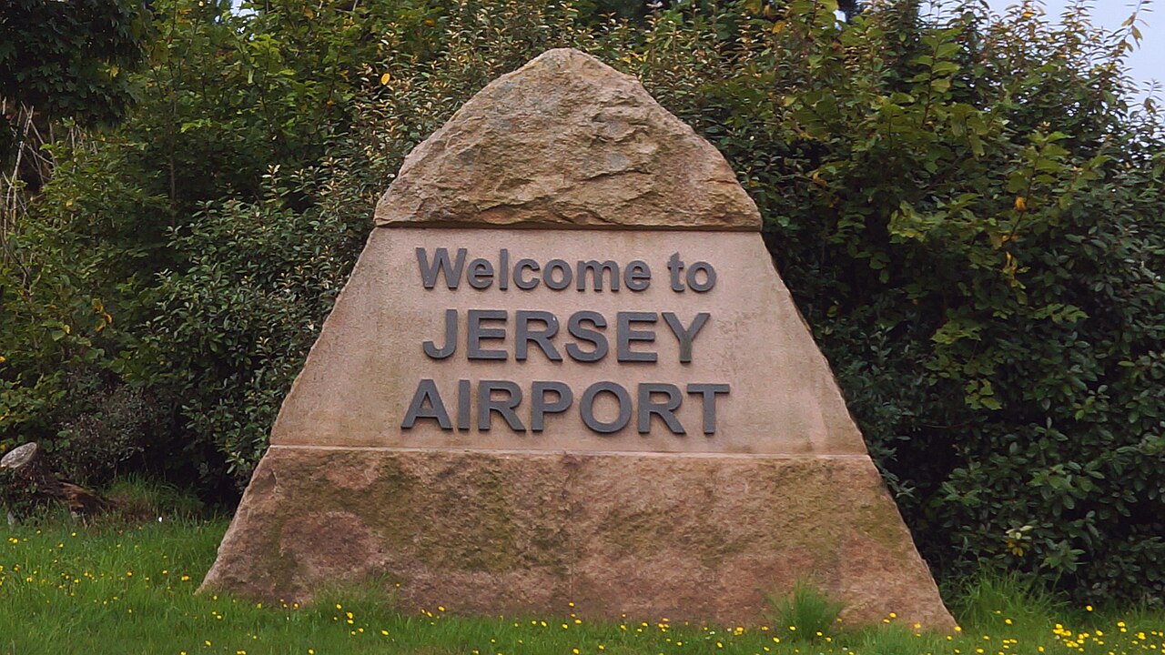 Rock sign that reads "Welcome to Jersey Airport" with grass, bushes, and shrubs in the background