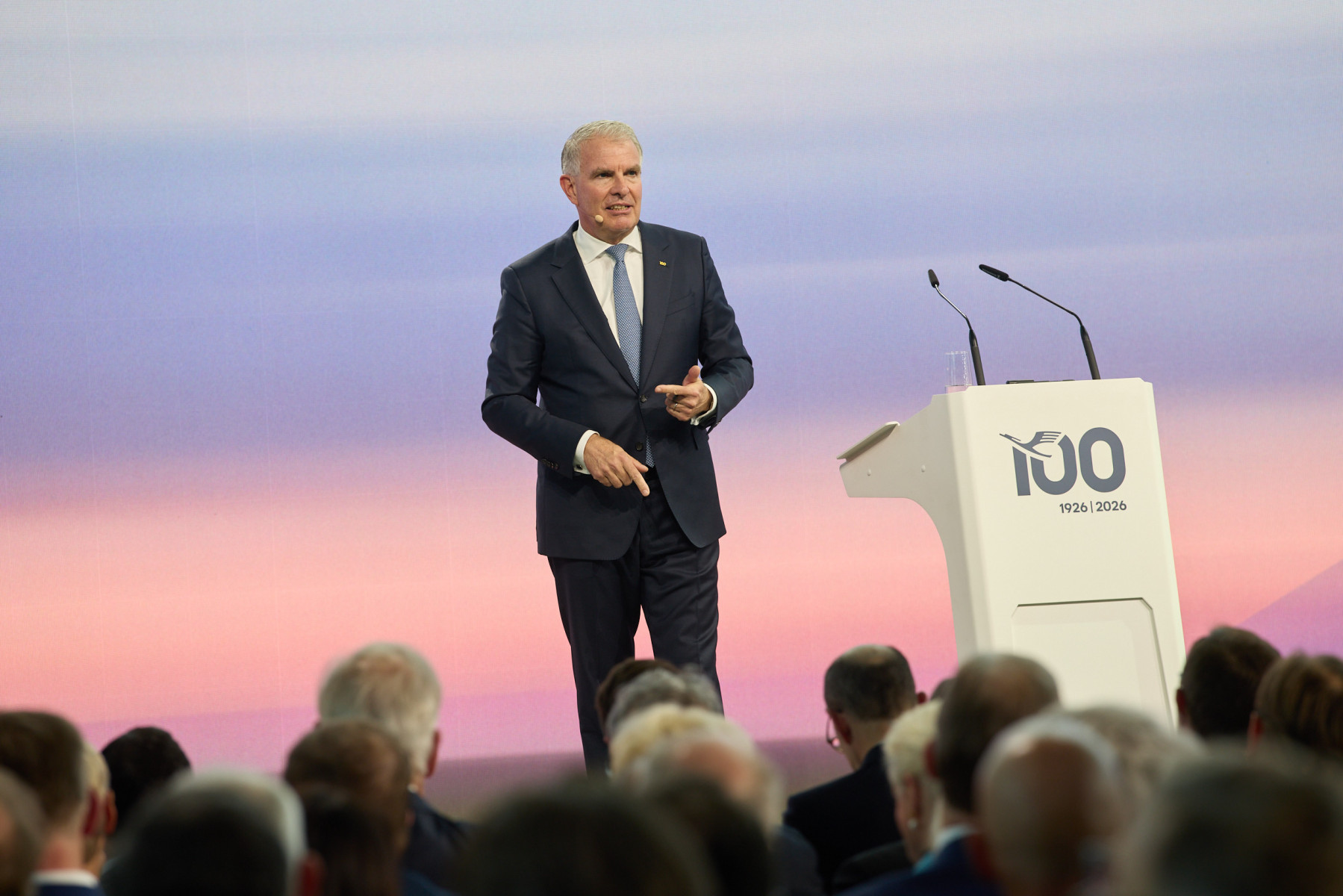 Carsten Spohr, CEO of Lufthansa Group standing by a white podium against a blue and pink background. Audience members can be seen at the bottom of the image.