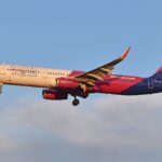 The image depicts a Wizz Air plane in flight. The aircraft is positioned in the centre of the image, with the nose facing towards the left, and has the pink and purpe branding of Wizz Air on the back end and tail. The sky is blue in the background, with some cloud beneath the plane along the bottom of the image.