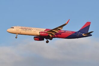 The image depicts a Wizz Air plane in flight. The aircraft is positioned in the centre of the image, with the nose facing towards the left, and has the pink and purpe branding of Wizz Air on the back end and tail. The sky is blue in the background, with some cloud beneath the plane along the bottom of the image.