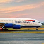 The front section of a British Airways plane on an airport taxiway against a pink and blue sky. The ground the plan is upon is dark grey concrete with yellow markings. The British Airways branding can be seen on the side of the plane's white body, and the engines are blue.