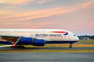 The front section of a British Airways plane on an airport taxiway against a pink and blue sky. The ground the plan is upon is dark grey concrete with yellow markings. The British Airways branding can be seen on the side of the plane's white body, and the engines are blue.
