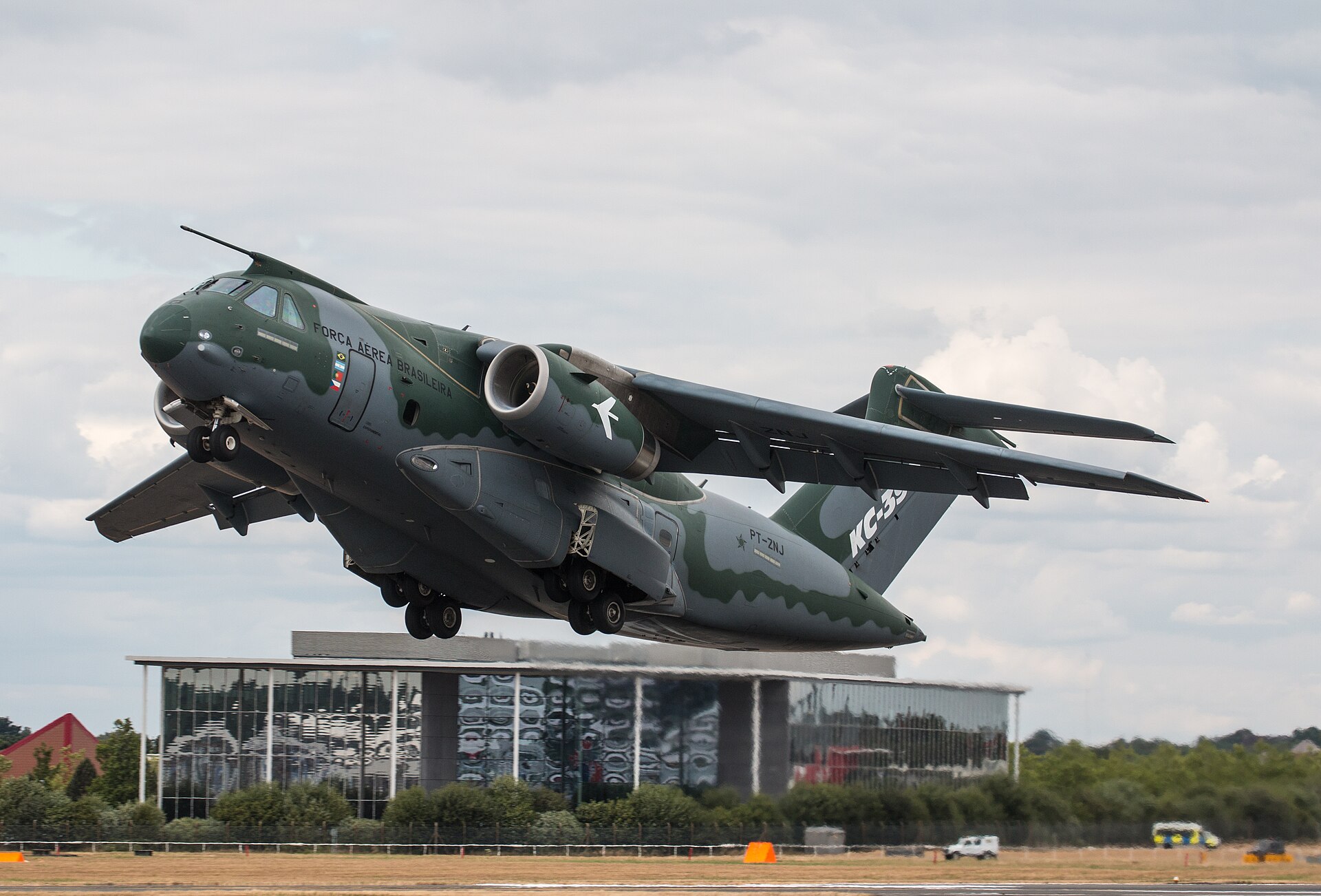 The image depicts the KC-390 Millennium flying low to the ground in take-off at Farnborough Air show. It has an army style dark green and grey camouflage pattern all over its body and wings. The sky is overcast in the background, and there is a small modern building behind in the mid distance.