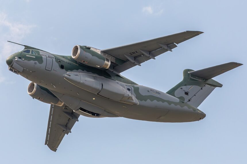 The image depicts an Embraer KC-390 aircraft in a blue sky from directly below