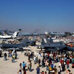People walk by and gather around a variation of aircraft at the International Air and Space fair in Chile. The image is taken from above. It is a nice day with a blue sky in the background