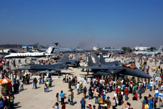 People walk by and gather around a variation of aircraft at the International Air and Space fair in Chile. The image is taken from above. It is a nice day with a blue sky in the background