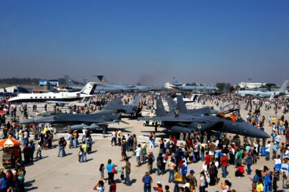 People walk by and gather around a variation of aircraft at the International Air and Space fair in Chile. The image is taken from above. It is a nice day with a blue sky in the background