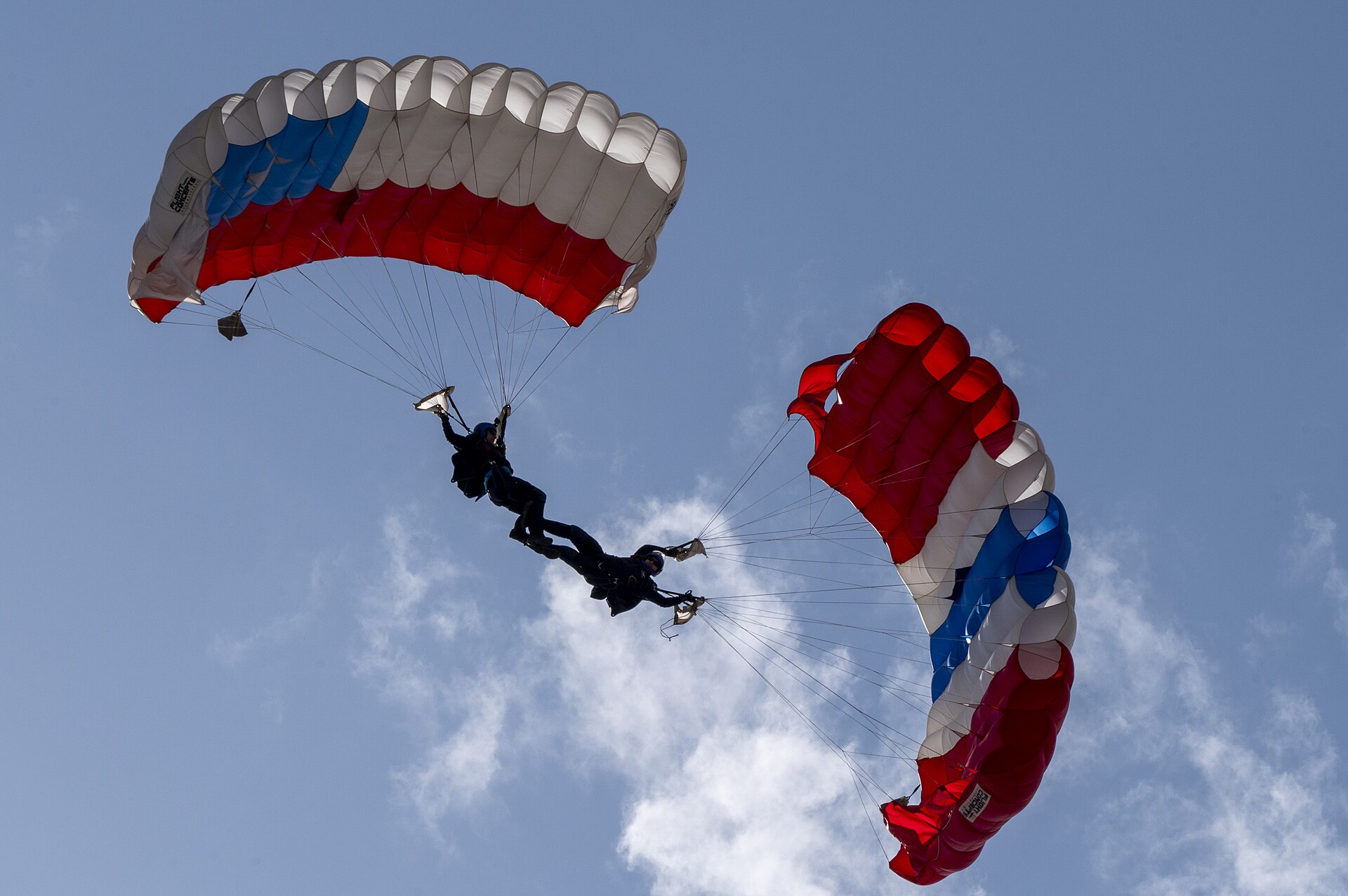 Two team members of the Chilean air force’s Boinas Azules (Blue Berets) Parachute Squadron drop during the Feria Internacional del Aire y del Espacio (FIDAE) at Pudahuel Air Base, Chile, April 13, 2024. The image is from beneath the parachuters, capturing the blue sky and their shadowy forms between two red blue and white parachutes.