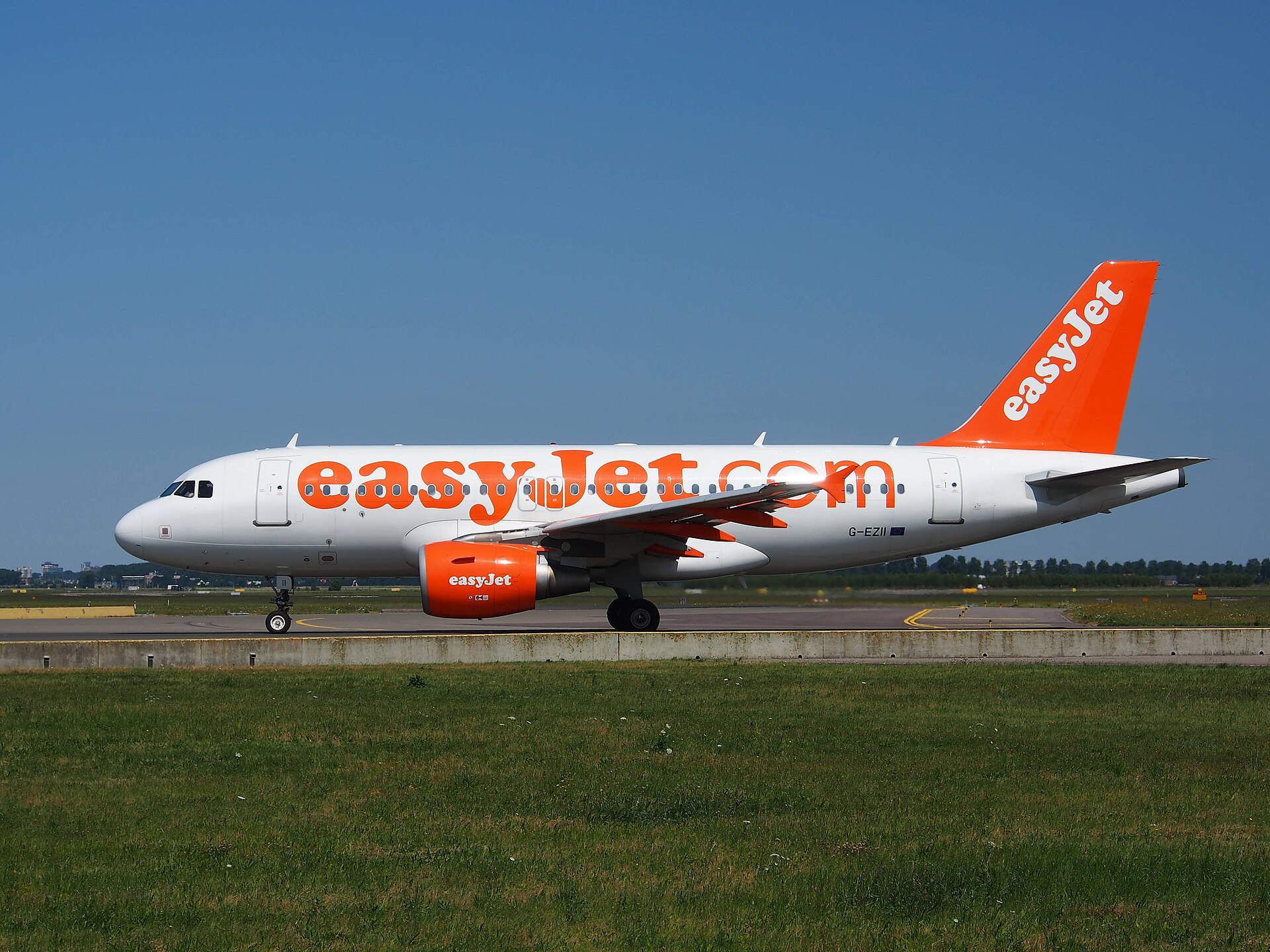 A white EasyJet plane with iconic orange branding on the body, jets, and tail is stationed near an airport runway. The grass is green in the foreground and the sky blue and cloudless in the background. The plane is central in the image, with the nose of the plane facing left. 