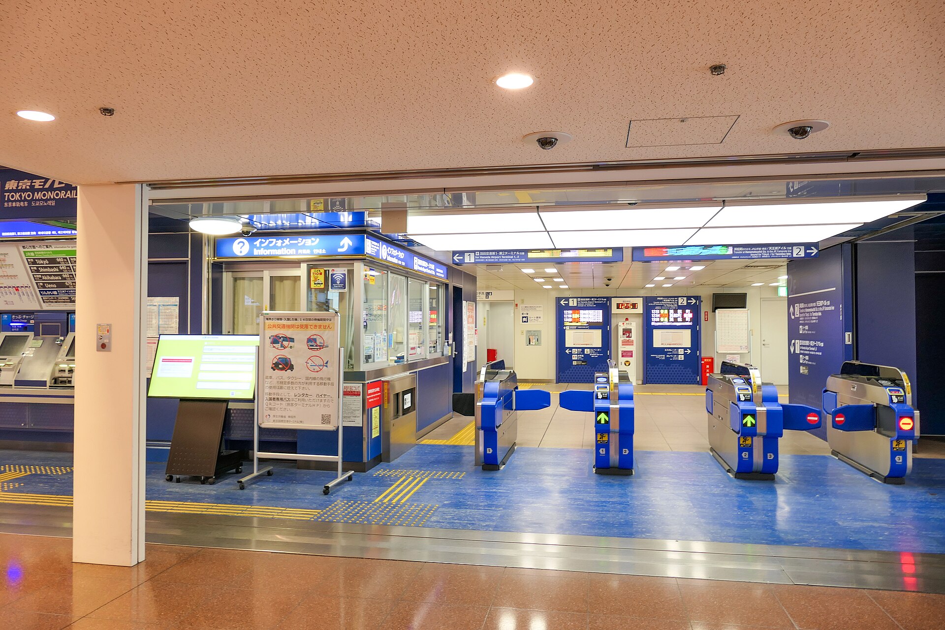 The ticket gate at Haneda Airport in Tokyo. The signs, scanners, floors and walls are cobalt blue. A help desk and ticket machine sit to the left of the gate. It is brightly lit and empty. 