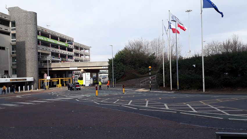 The image depicts the outside of Terminal 1 of Manchester airport on an overcast day. The Terminal building is in the upper left quadrant of the image, the lower half shows the tarmac drop-off point. The centre right of the image shows hedges and an overcast sky, as well as 3 flagpoles. 