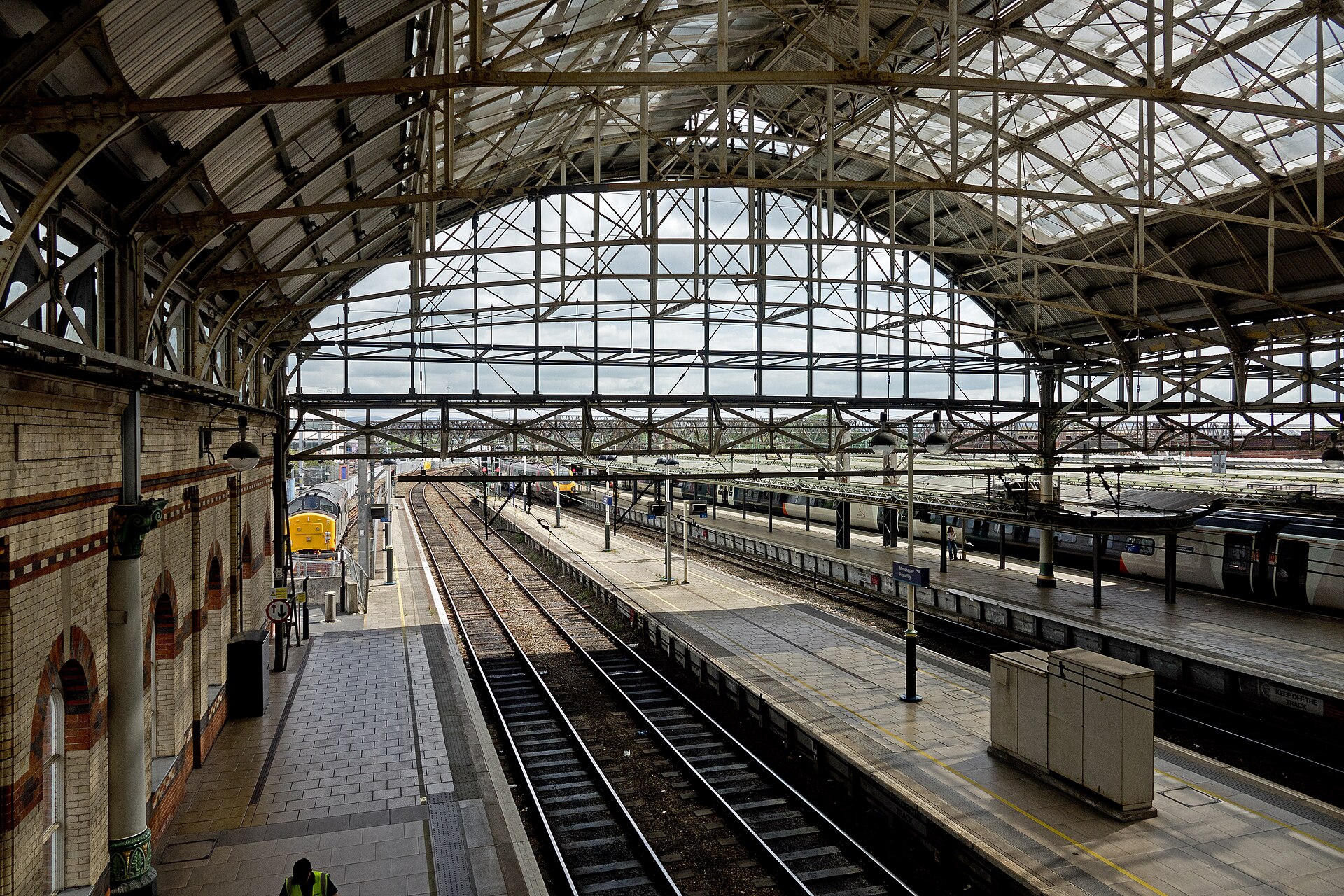 The image depicts the far end of two platforms at Manchester Piccadilly Station, taken from above looking down and out. The main focus of the image is the structure of the arched beams across the ceiling, which take up the image from the top to the centre. The lower half of the image focuses on the train tracks as they extend out of the station. 