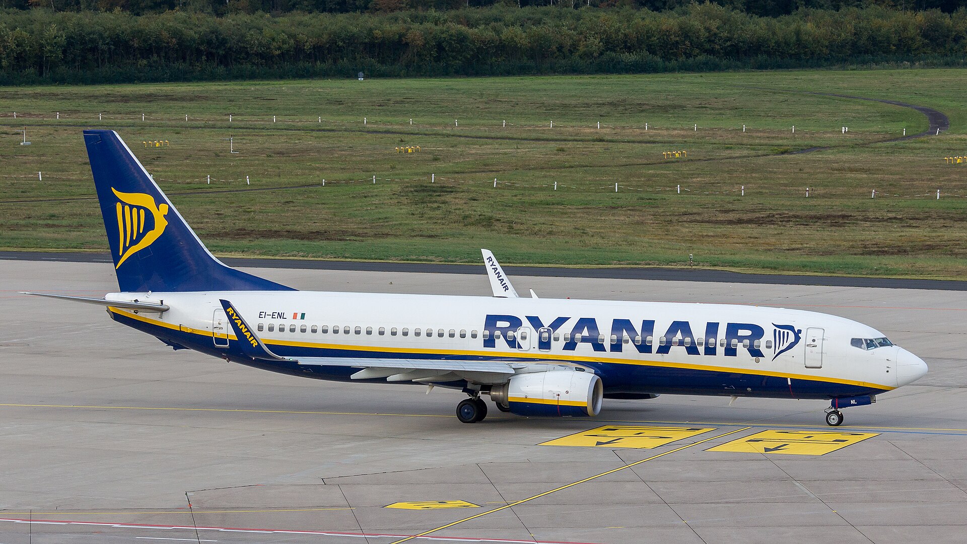 A Ryanair plane on the ground in an airport taxiway. It is in the centre of the image, the nose facing the right and the tail on the left. The plane is white with the blue and yellow Ryanair logo on the tail, with 'Ryanair' written in dark blue across the body of the plane. The background is grass. 