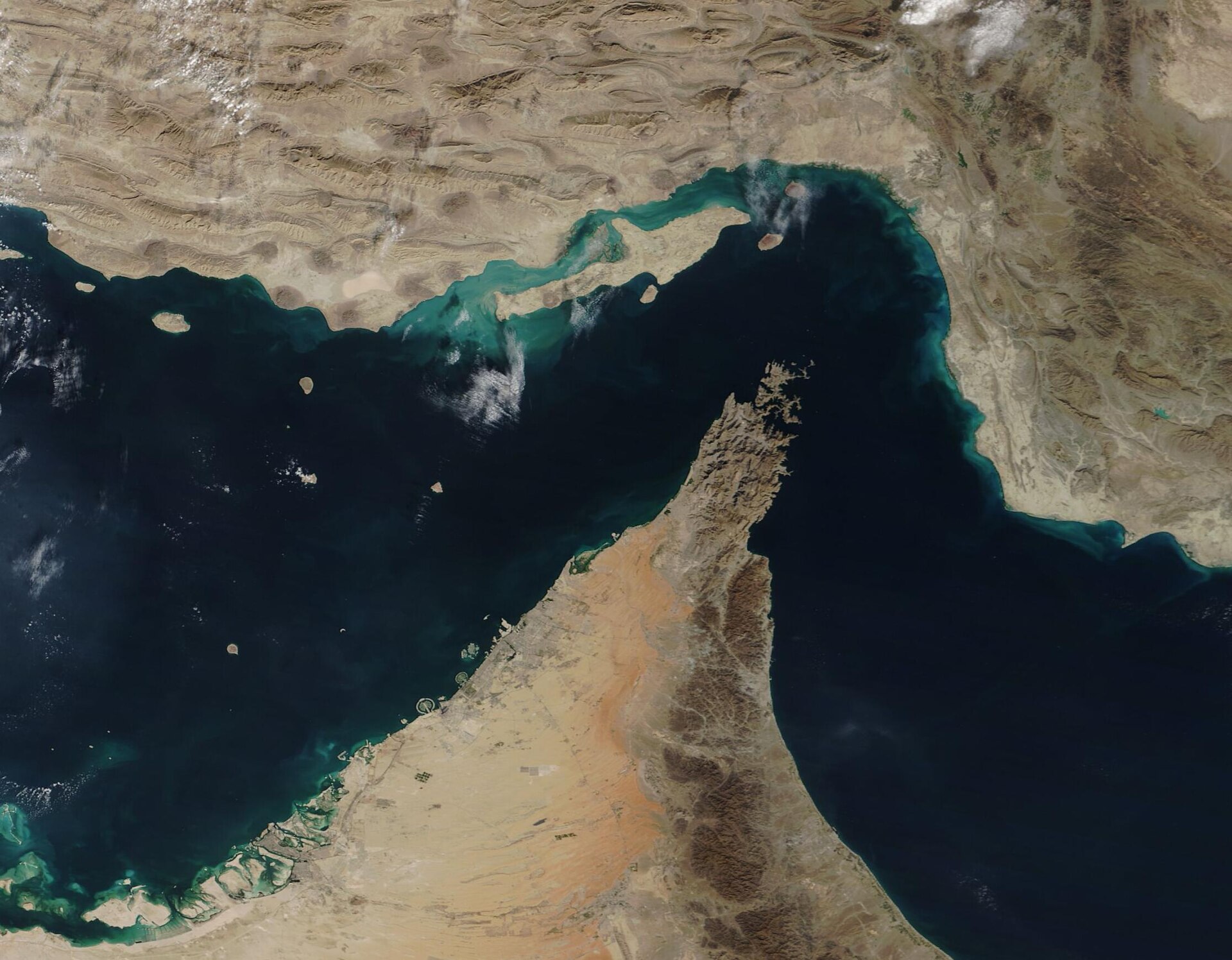 The image depicts an aerial view of the Straight of Hormuz. The body of water which goes across the image is a deep blue-green, and the land around it at the top and bottom of the image is various shades of beige and brown. 