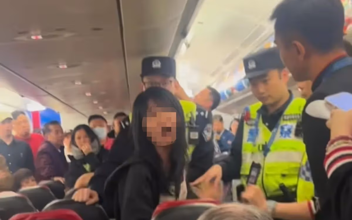 Women with blurred face looking at the camera. Cabin crew and local authorities surrounding her. Other passengers seen stood up in the background watching her.