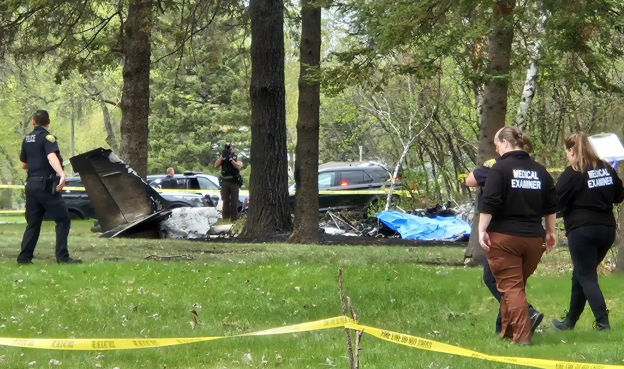 In a field, area with police tape. Medical examiners on scene, plane crash among trees on the floor in the background. Forensics taking photos, police cars in the background.