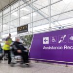 London Stansted Airport interior, with large glass windows and a purple sign with the words disability reception on them. A person pushes another person in a wheelchair.