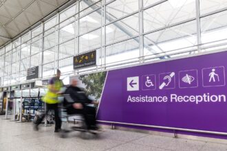 London Stansted Airport interior, with large glass windows and a purple sign with the words disability reception on them. A person pushes another person in a wheelchair.