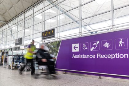 London Stansted Airport interior, with large glass windows and a purple sign with the words disability reception on them. A person pushes another person in a wheelchair.