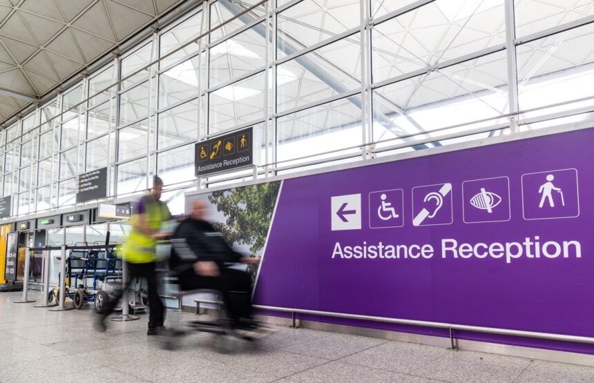 London Stansted Airport interior, with large glass windows and a purple sign with the words disability reception on them. A person pushes another person in a wheelchair.