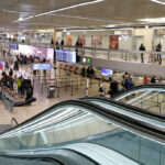 Geneva airport queues at border. Escalators in view, crowded room.