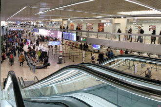 Geneva airport queues at border. Escalators in view, crowded room.