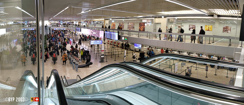 Geneva airport queues at border. Escalators in view, crowded room.
