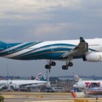 An Oman Air aircraft landing in Heathrow Airport. The aircraft is blue and white and is landing on a gravelly track among other aircrafts.
