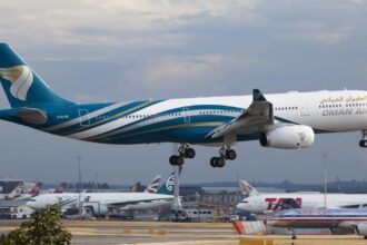 An Oman Air aircraft landing in Heathrow Airport. The aircraft is blue and white and is landing on a gravelly track among other aircrafts.