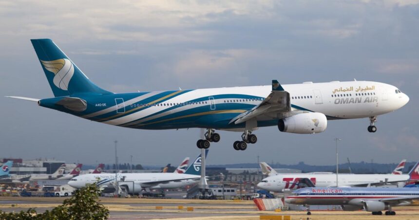 An Oman Air aircraft landing in Heathrow Airport. The aircraft is blue and white and is landing on a gravelly track among other aircrafts.