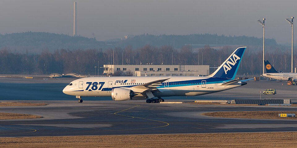 Boeing 787-8 Dreamliner reg. JA814A, on the runway. Blue and white livery. Trees and building in the background.