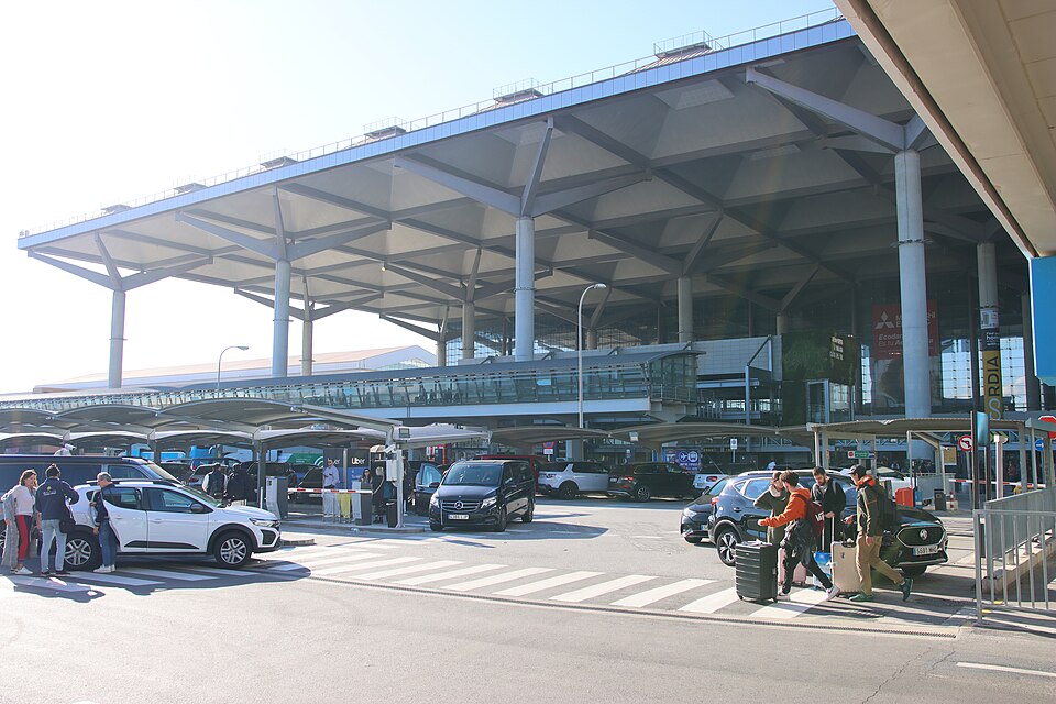 Image shows the exterior of Málaga airport on a sunny day with cars parked outside and people with suitcases