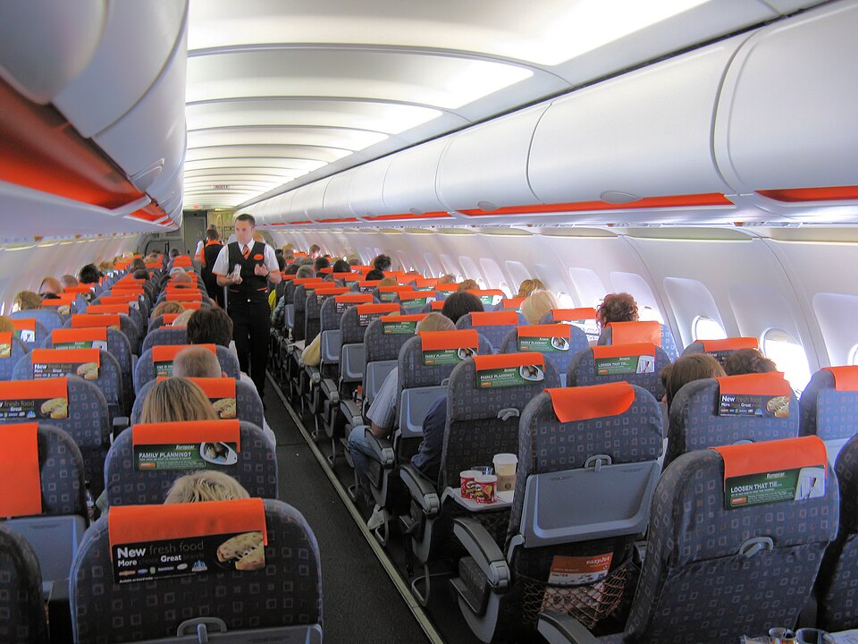 An interior view of Easy Jet’s airplane cabin. Cabin crew stood up walking down isle, passengers in seat.