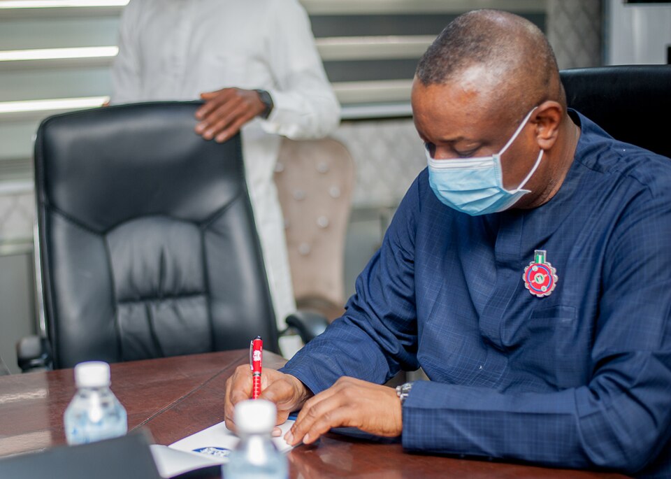 A photograph of Festus Keyamo wearing a blue surgical mask and a dark blue traditional outfit while signing documents during a 2020 visit to Mojec International.