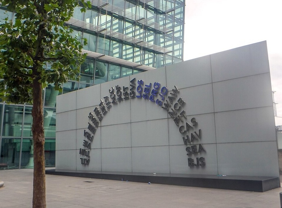 The image depicts an arched sign of airport IATA codes outside London Heathrow Airport. An airport building stands behind with floor to ceiling glass windows. A tree is in the front left of the image.
