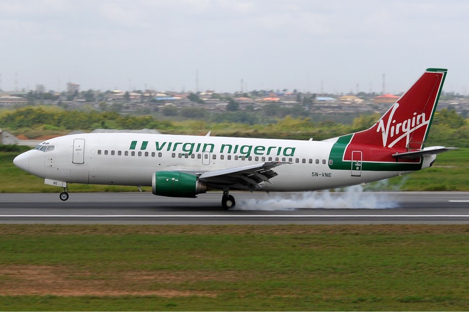 A Virgin Nigeria Boeing 737-300 aircraft touching down on a runway, with a puff of smoke visible from the tires upon landing.