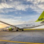 An A220-300 aircraft is in a taxiway, the tail has an airBaltic logo on it, and is closest to the camera. The sun is in the distance (the bottom left of the image) as stairs descend from the plane's main door.