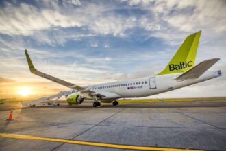 An A220-300 aircraft is in a taxiway, the tail has an airBaltic logo on it, and is closest to the camera. The sun is in the distance (the bottom left of the image) as stairs descend from the plane's main door.