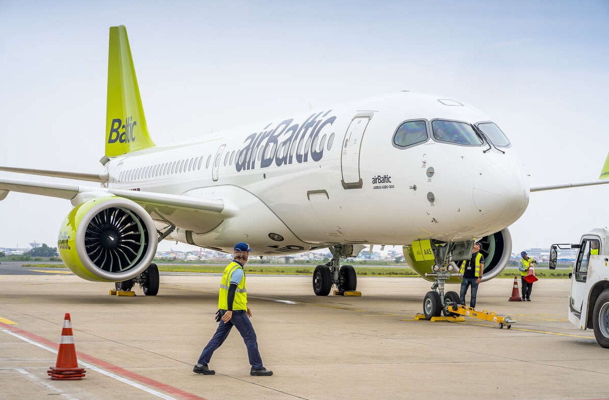 An airBaltic fleet Airbus A220-300 in an airport taxiway, the front facing the camera at an angle. The signature pale green tail can be seen in the top left corner f the image, and dark blue 'airBaltic' logo can be seen on the side of the plane. A man in high-vis walks in front of the plane facing away from the camera