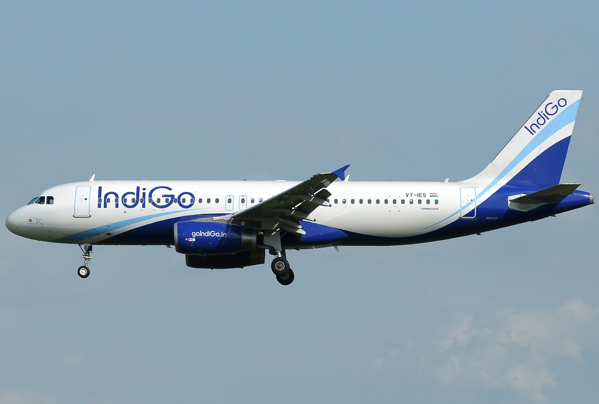 A white and blue IndiGo passenger airplane captured mid-flight against a clear sky, viewed from the side.