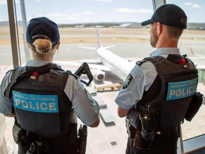 Two Australian Federal police officers in the foreground look out of an upper window of an airport to a plane in the taxiway 