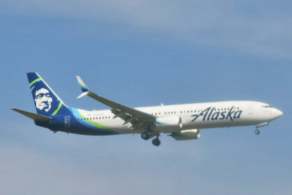 An Alaska Airlines passenger jet flies through a hazy gray sky with its landing gear extended as it approaches for landing
