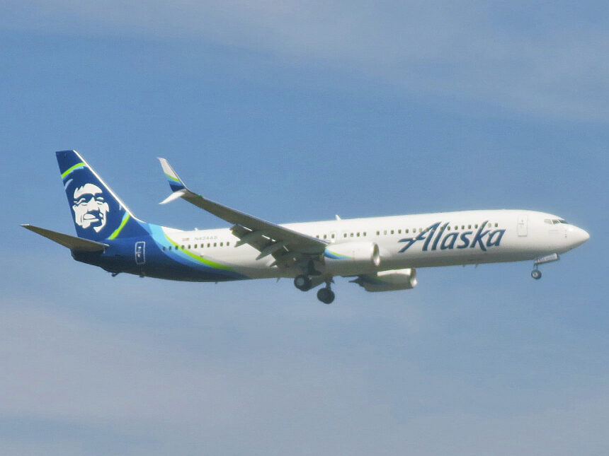 An Alaska Airlines passenger jet flies through a hazy gray sky with its landing gear extended as it approaches for landing
