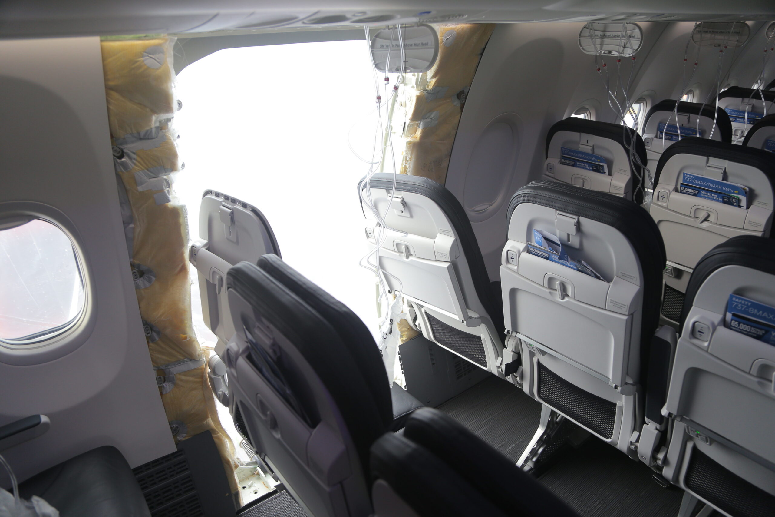 Interior view of a narrow‑body aircraft cabin showing a row of grey passenger seats and oval windows on the right side, with seatback trays, safety cards and mesh pockets visible. On the left side, a large section of the fuselage wall is missing, revealing a bright opening to the outside, with exposed yellow insulation, wiring and structural panels around the edges. Loose cables and oxygen lines hang from the ceiling above the opening, contrasting with the otherwise intact seating and cabin fittings.