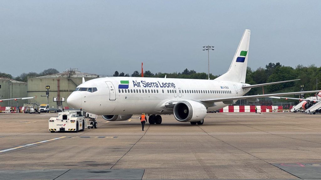 Side view of a white Air Sierra Leone passenger jet parked on an airport apron, with the airline name and green-and-blue tail markings clearly visible on the fuselage. The aircraft is positioned nose‑on towards the camera, connected to a white pushback tug at the front landing gear. A ground crew member in high‑visibility clothing walks across the concrete apron in front of the aircraft. In the background, low industrial airport buildings, red‑and‑white safety barriers, service vehicles and trees line the edge of the airfield beneath a grey, overcast sky.
