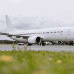 Side view of a white, unbranded narrow‑body passenger aircraft parked on an airport apron, photographed from a low angle across a strip of green grass in the foreground. The aircraft’s fuselage, wings and tail are clean and undecorated, with engines mounted beneath the wings and the landing gear resting on the concrete surface. Ground service equipment and mobile stairs are positioned near the forward section, while low airport buildings and trees sit in the background. The scene is lit by soft daylight under a grey, cloud‑filled sky, giving the image a muted, industrial atmosphere.